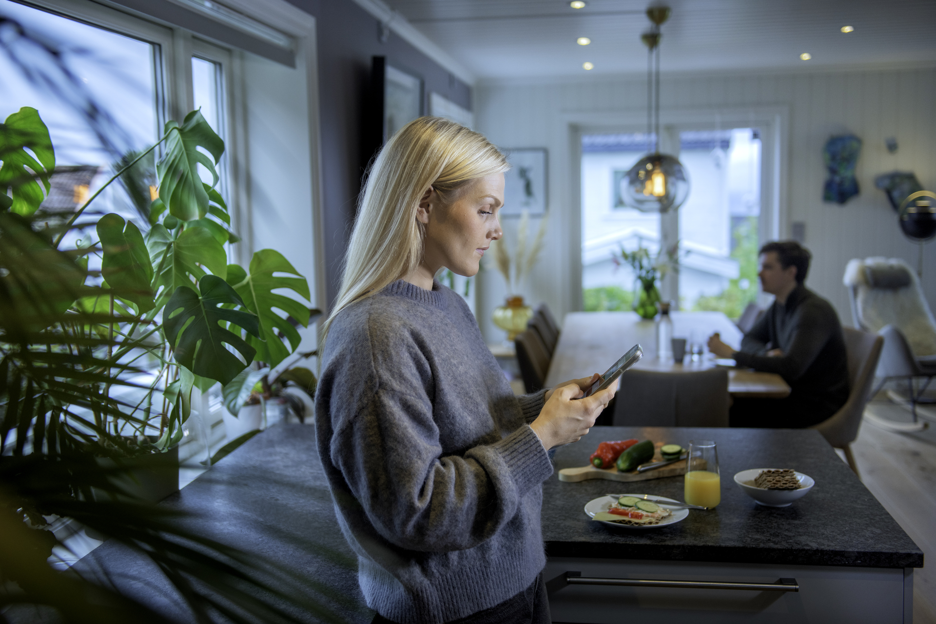 Kvinne som bekymret leser på telefon hjemme, en mann sitter ved spisebordet i bakgrunnen og ser ut i lufta.