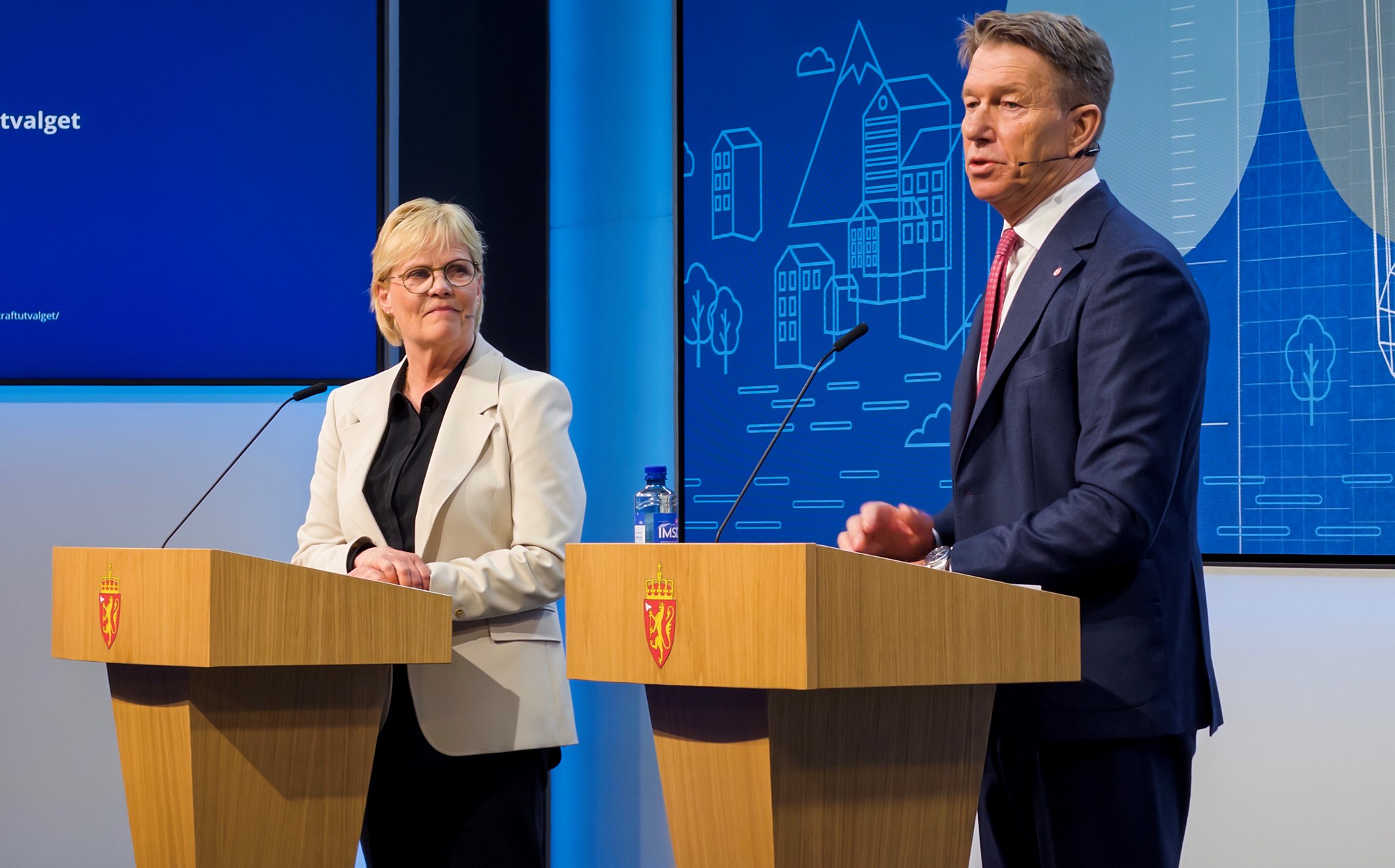 Two people stand at podiums with Norway's national coat of arms. A screen with illustrations of buildings appears in the background. Photo: Jo Henrik Jarstø/Ministry of Energy
