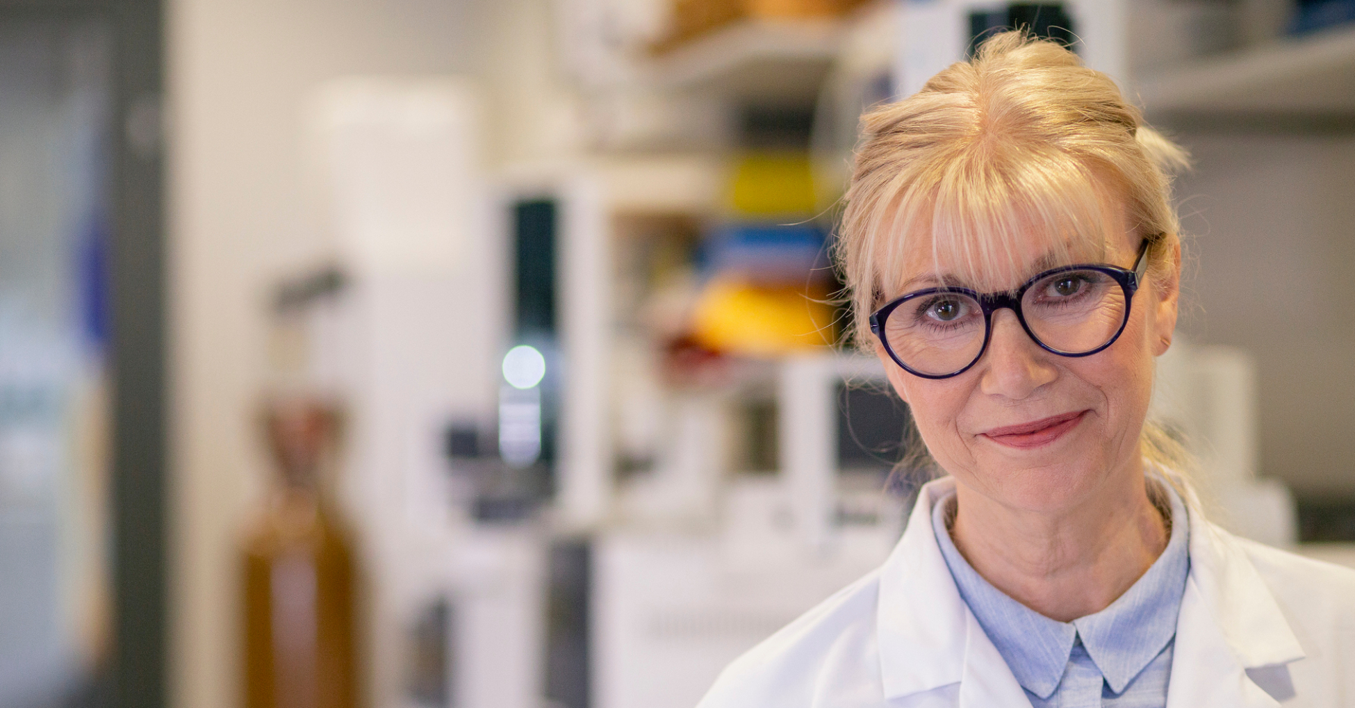 A woman in a white lab coat smiles kindly. She has blond hair, glasses and is standing in a modern laboratory. Photo: Getty Images
