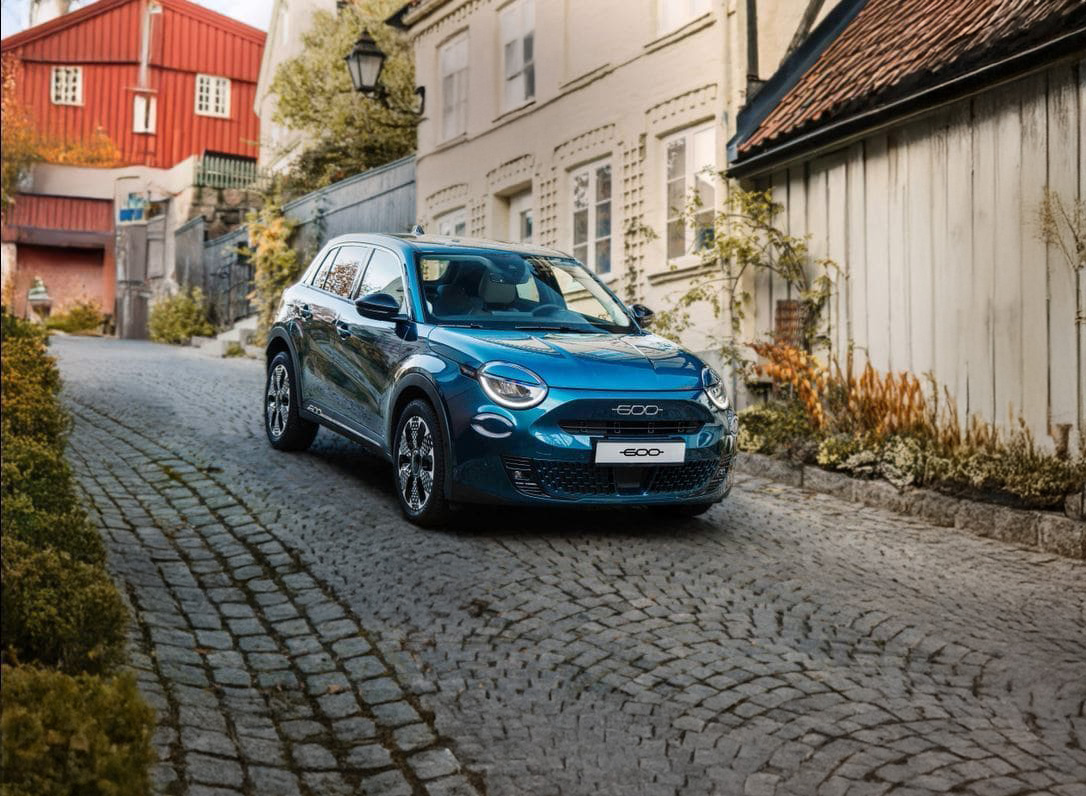 En blå Fiat 600 parkert i en brosteinsbelagt gate omgitt av trehus og høstfargede planter.