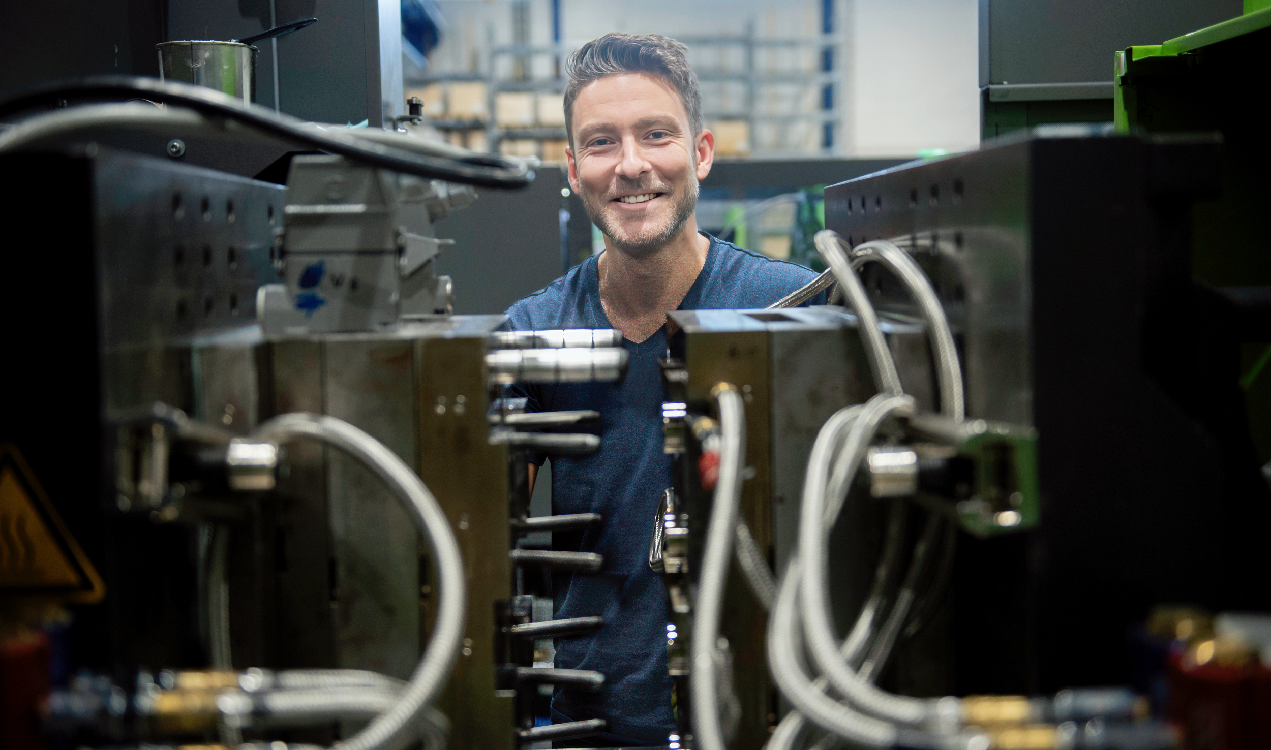 A person works at a large industrial machine surrounded by metal components and cables in a production environment.