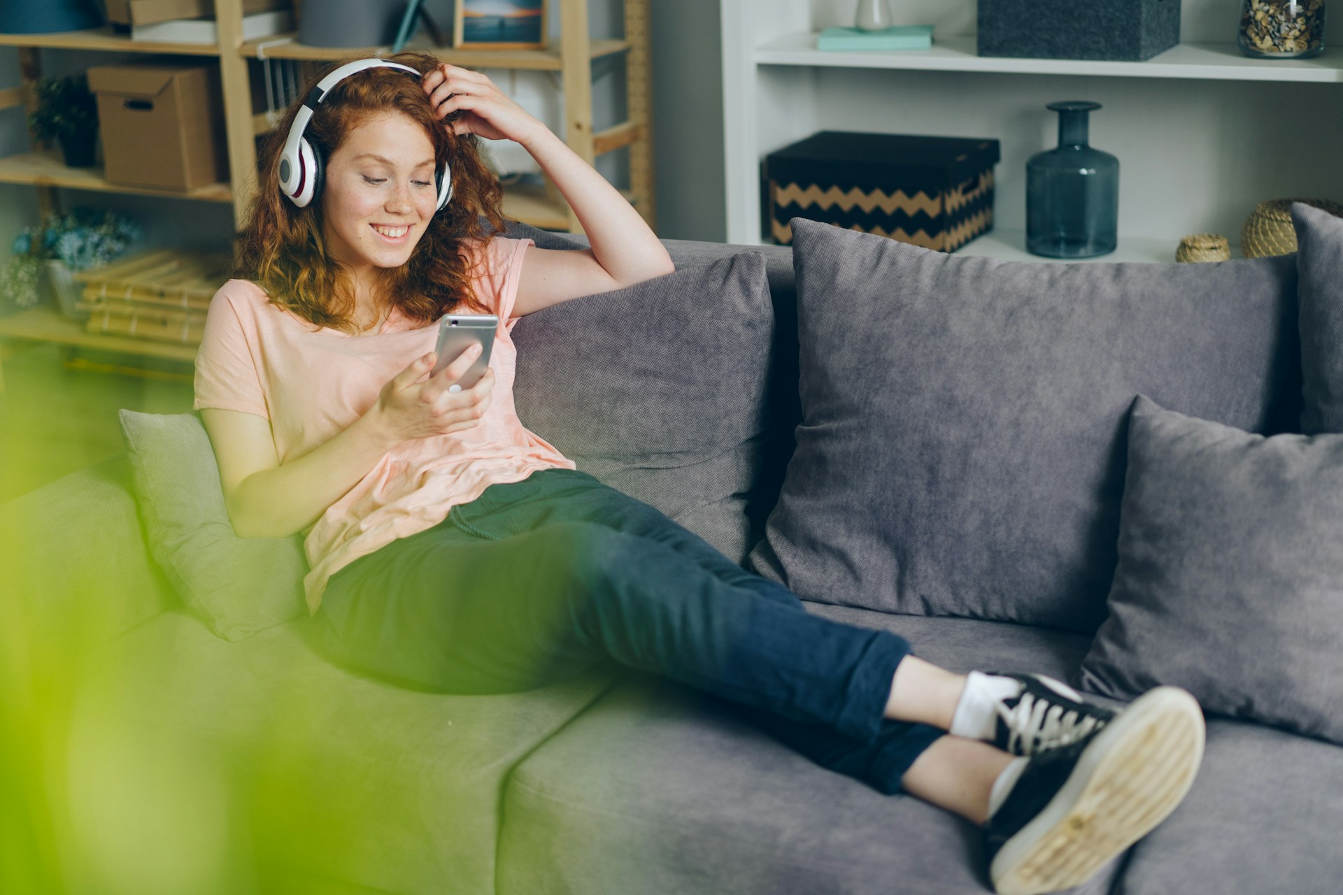 En smilende jente med hodetelefoner slapper av i sofaen hjemme og ser på mobiltelefonen.