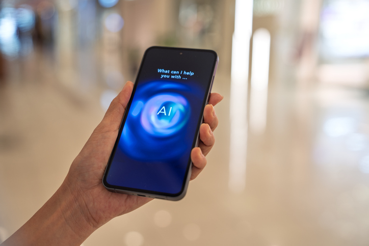 Hand holds a smartphone showing an AI assistant. On the screen is the text "What can I help you with..." above a blue circular graphic with the letters "AI".