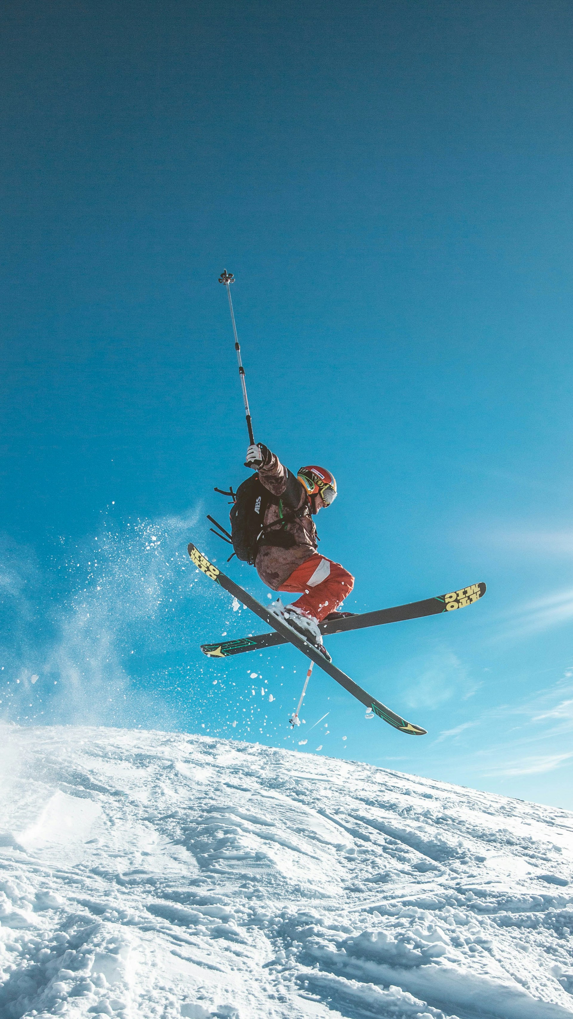 Freestyler på ski gjør et høyt hopp over en snødekt bakke, med skiene i kryss i luften og en stav løftet over hodet. Snøsprut står bak utøveren, og himmelen er klar og knallblå, noe som gir et dramatisk og energisk uttrykk.