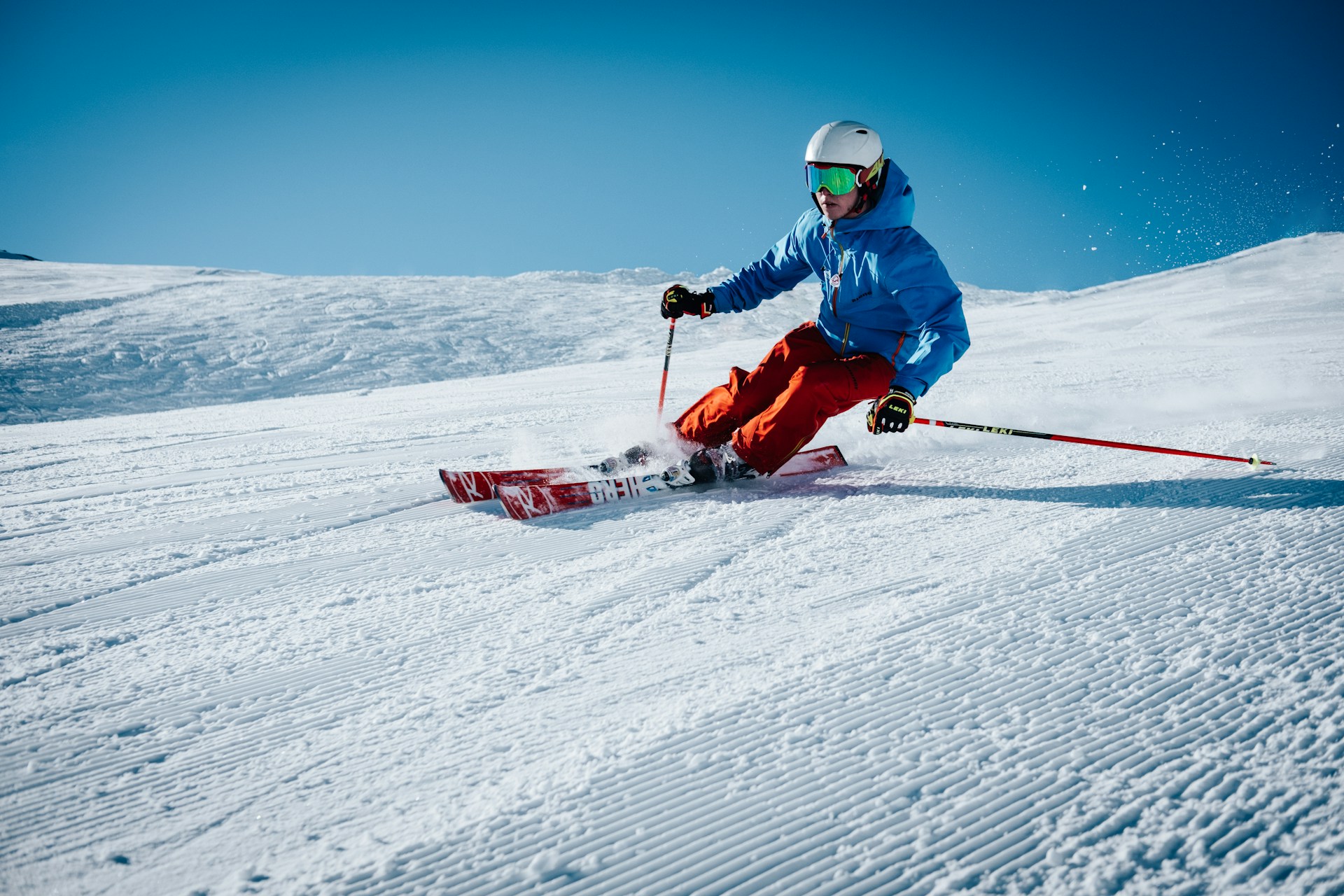 En person i blå jakke og røde bukser står på alpinski nedover en nypreparert, solfylt skibakke med klar,blå himmel i bakgrunnen. Snøspruten står rundt skiene.