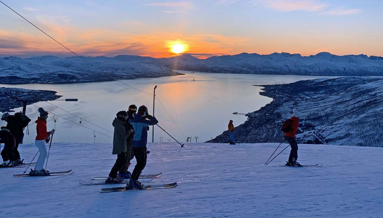 Bilde fra Tromsø Alpinpark. Flere personer på ski står og kjører rolig ned en snødekt fjellside mens sola går ned over en bred fjord omgitt av snødekte fjell. Vannflaten speiler det varme oransje lyset fra solnedgangen, og stemningen er rolig og vinterlig.