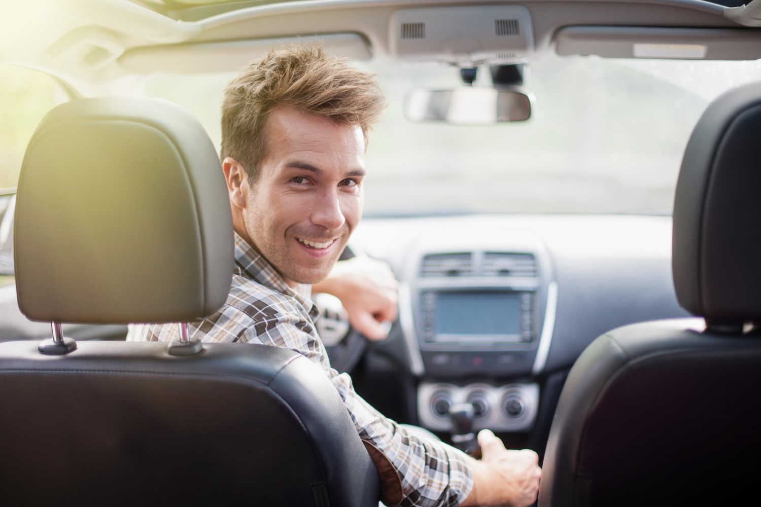 En mann sitter i førersetet i bilen og ser bakover mellom forsetene i et lyst bilinteriør. Mannen smiler.