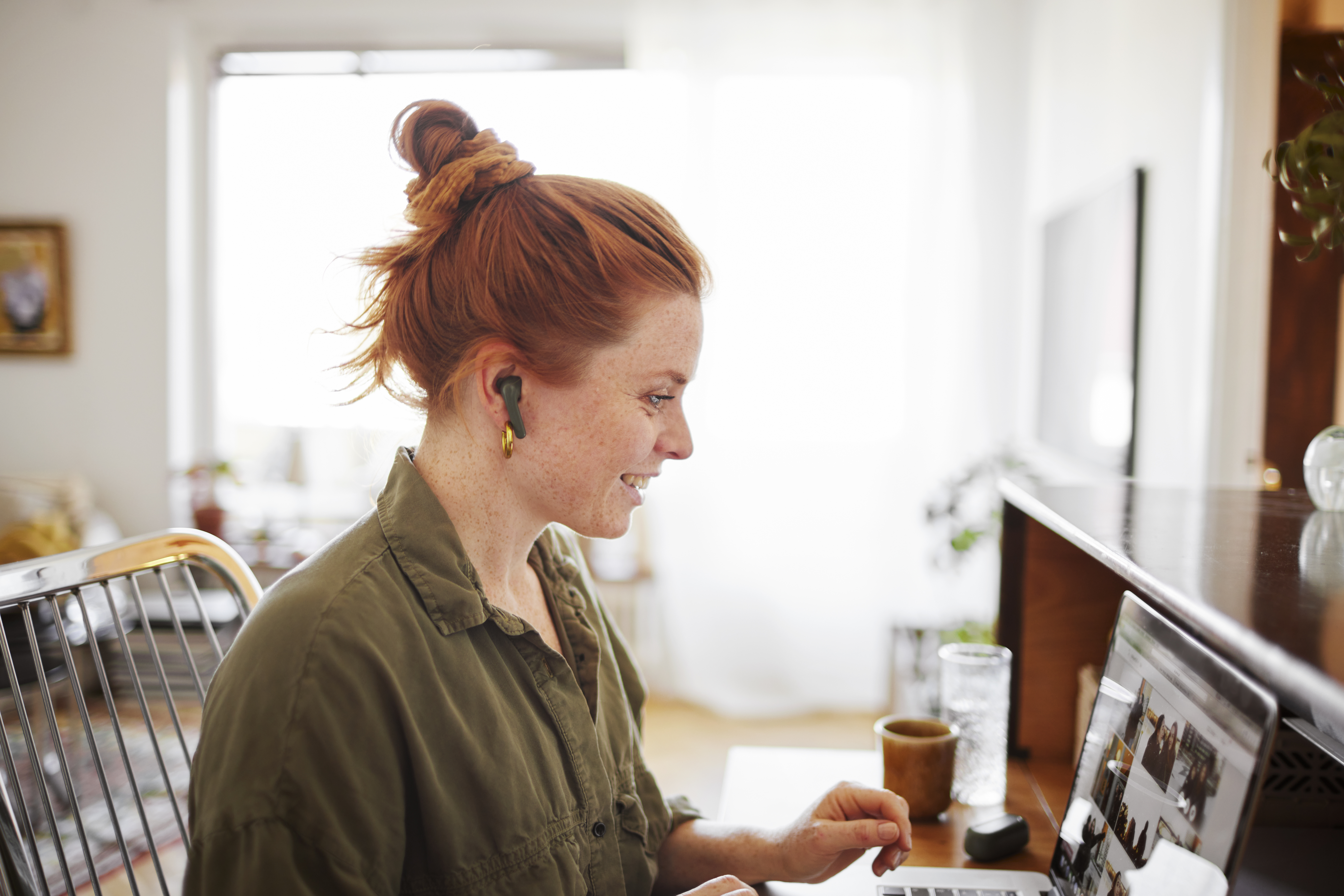 Woman with red hair on digital Intro 2 course, working on laptop at home office with coffee cup and glass on the table in a bright living room.